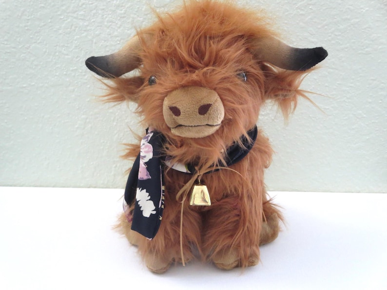K&ouml;nnte beinhalten: Ein Pl&uuml;schtier, das eine Highland-Kuh darstellt, mit zotteligem braunem Fell, schwarzen H&ouml;rnern und einem schwarzen Schal mit Blumenmuster. Es hat eine kleine goldene Glocke um den Hals. Das Spielzeug steht auf einer wei&szlig;en Oberfl&auml;che.