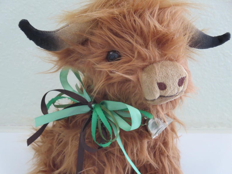 K&ouml;nnte beinhalten: Ein Pl&uuml;sch-Highland-Kuh mit zotteligem, hellbraunem Fell, schwarzen H&ouml;rnern und einer hellbraunen Schnauze. Es hat ein gr&uuml;nes und braunes Band um den Hals gebunden. Das Spielzeug hat einen freundlichen Ausdruck.