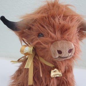 K&ouml;nnte beinhalten: Ein Pl&uuml;sch-Highland-Rind mit zotteligem, rotbraunem Fell. Es hat schwarz gef&auml;rbte H&ouml;rner, eine hellbraune Schnauze und eine goldene Glocke um den Hals. Ein goldenes Band ist um den Hals gebunden und verleiht ihm eine dekorative Note. Das Rind ist ein Stofftier.