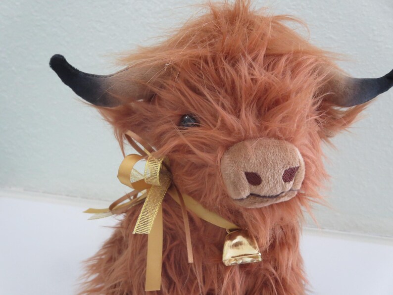 K&ouml;nnte beinhalten: Ein Pl&uuml;sch-Highland-Rind mit zotteligem, rotbraunem Fell. Es hat schwarz gef&auml;rbte H&ouml;rner, eine hellbraune Schnauze und eine goldene Glocke um den Hals. Ein goldenes Band ist um den Hals gebunden und verleiht ihm eine dekorative Note. Das Rind ist ein Stofftier.