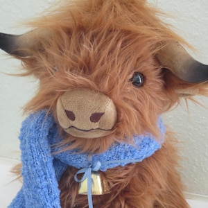 K&ouml;nnte beinhalten: Ein Stofftier, das eine Highland-Kuh darstellt, mit langem, zotteligem braunem Fell und einer hellbraunen Schnauze. Es hat dunkle H&ouml;rner und tr&auml;gt einen blauen Strickschal und eine kleine goldene Glocke. Die Kuh ist etwa 25 cm gro&szlig;.