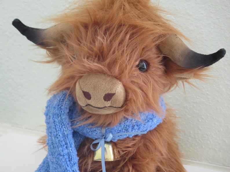 K&ouml;nnte beinhalten: Ein Stofftier, das eine Highland-Kuh darstellt, mit langem, zotteligem braunem Fell und einer hellbraunen Schnauze. Es hat dunkle H&ouml;rner und tr&auml;gt einen blauen Strickschal und eine kleine goldene Glocke. Die Kuh ist etwa 25 cm gro&szlig;.