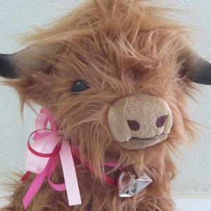 K&ouml;nnte beinhalten: Ein Pl&uuml;sch-Highland-Rind mit zotteligem, braunem Fell, schwarzen H&ouml;rnern und einem rosa Band um den Hals. Es hat eine weiche, beige Schnauze und eine silberne Glocke. Das Spielzeug ist einem Highland-Rind nachempfunden.