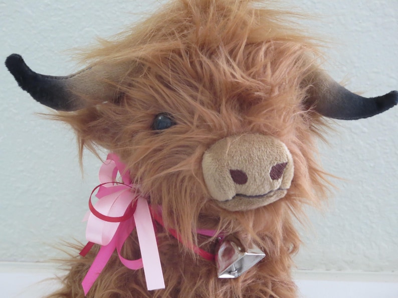 K&ouml;nnte beinhalten: Ein Pl&uuml;sch-Highland-Rind mit zotteligem, braunem Fell, schwarzen H&ouml;rnern und einem rosa Band um den Hals. Es hat eine weiche, beige Schnauze und eine silberne Glocke. Das Spielzeug ist einem Highland-Rind nachempfunden.