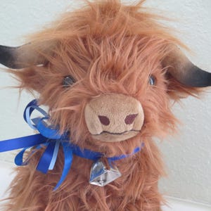 K&ouml;nnte beinhalten: Ein Pl&uuml;sch-Highland-Rind mit zotteligem, rotbraunem Fell, schwarz gef&auml;rbten H&ouml;rnern und einer hellbraunen Schnauze. Es tr&auml;gt ein blaues Bandhalsband mit einer silbernen Glocke. Das Spielzeug hat einen freundlichen Ausdruck.