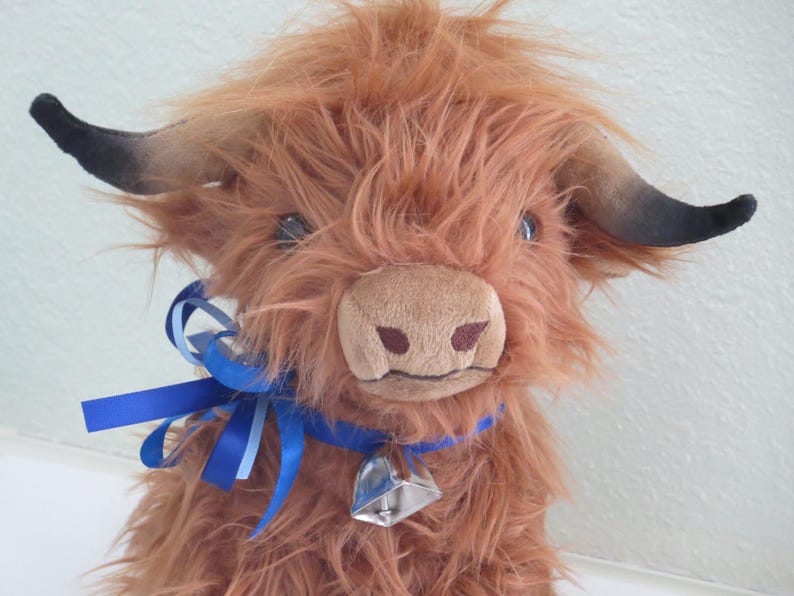 K&ouml;nnte beinhalten: Ein Pl&uuml;sch-Highland-Rind mit zotteligem, rotbraunem Fell, schwarz gef&auml;rbten H&ouml;rnern und einer hellbraunen Schnauze. Es tr&auml;gt ein blaues Bandhalsband mit einer silbernen Glocke. Das Spielzeug hat einen freundlichen Ausdruck.