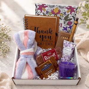 Puede incluir: Una caja de regalo llena de varios artículos. Incluye una bata suave a rayas, un cuaderno con el texto "Thank you", una vela, chocolate, galletas y un bolígrafo. La caja está decorada con motivos florales y contiene productos con aroma a lavanda.