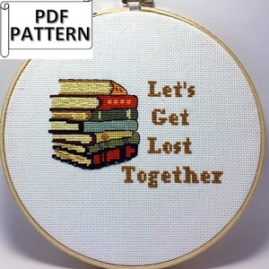 May include: A white embroidery hoop with a brown wooden frame. The hoop contains a cross-stitch design of a stack of books with the text "Let's Get Lost Together" in brown thread.