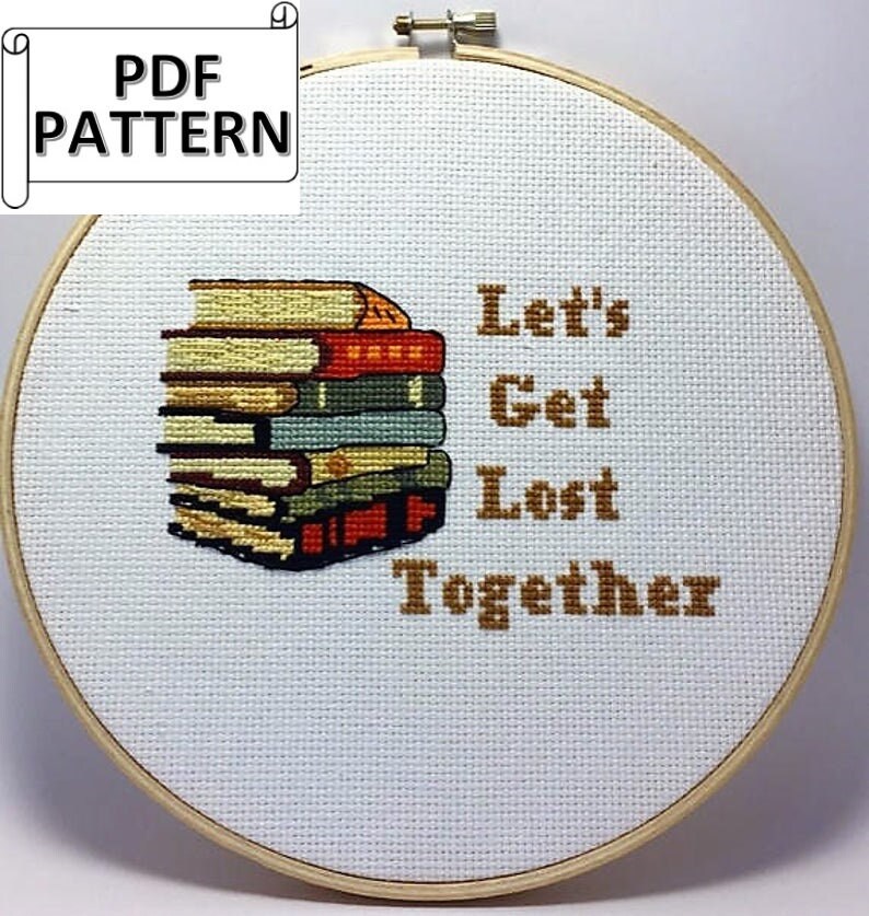 May include: A white embroidery hoop with a brown wooden frame. The hoop contains a cross-stitch design of a stack of books with the text "Let's Get Lost Together" in brown thread.