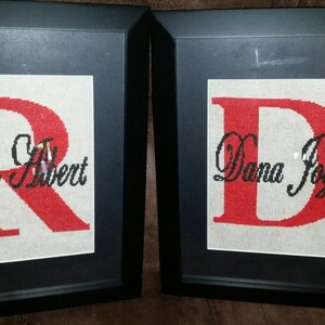 May include: Two framed cross-stitch pieces with the letters "R" and "D" in red. The "R" has the name "Roy Albert" written in cursive below it. The "D" has the name "Dana Joyce" written in cursive below it.
