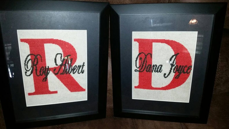 May include: Two framed cross-stitch pieces with the letters "R" and "D" in red. The "R" has the name "Roy Albert" written in cursive below it. The "D" has the name "Dana Joyce" written in cursive below it.