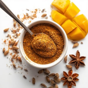 May include: A white bowl filled with a brown spice blend, with a spoon resting inside. Cubed mango and star anise are arranged around the bowl. Cardamom pods and spice granules are scattered on a white surface, creating a culinary still life.