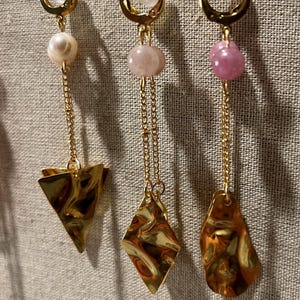 May include: Three pairs of gold-toned earrings with different designs. Each earring features a clear, round stud, a chain, and a unique charm: a pearl and triangle, a pink stone and diamond, and a pink stone and teardrop shape. The earrings hang against a neutral fabric.
