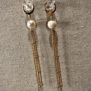 May include: A pair of gold-coloured drop earrings. Each earring has a clear stud, a gold hoop, a white pearl, and multiple strands of delicate gold chains. The earrings are set against a neutral, textured background.