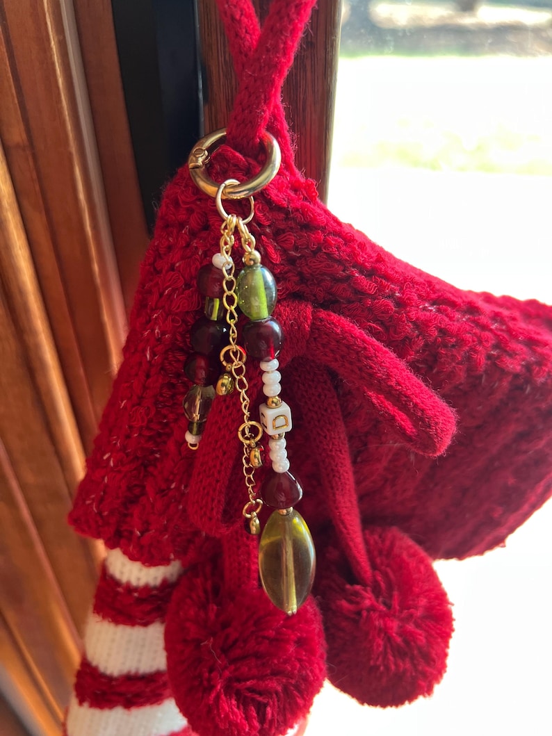 K&ouml;nnte beinhalten: Ein roter gestrickter Weihnachtsstrumpf mit wei&szlig;en Streifen und einem goldfarbenen Ring, der einen Perlenanh&auml;nger h&auml;lt. Der Anh&auml;nger hat gr&uuml;ne, rote und wei&szlig;e Perlen und eine kleine goldfarbene Kette.