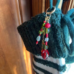 K&ouml;nnte beinhalten: Eine gr&uuml;ne Stricktasche mit einem goldfarbenen Verschluss und einem Perlenanh&auml;nger. Der Anh&auml;nger hat rote, gr&uuml;ne und klare Perlen, sowie einen facettierten roten Kristallanh&auml;nger und einen kleinen goldfarbenen Buchstaben "H".
