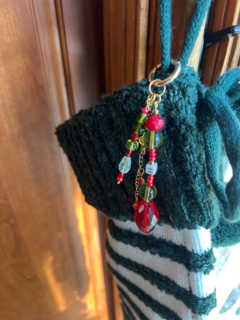 K&ouml;nnte beinhalten: Eine gr&uuml;ne Stricktasche mit einem goldfarbenen Verschluss und einem Perlenanh&auml;nger. Der Anh&auml;nger hat rote, gr&uuml;ne und klare Perlen, sowie einen facettierten roten Kristallanh&auml;nger und einen kleinen goldfarbenen Buchstaben "H".