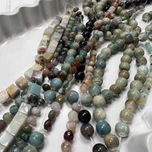 Puede incluir: Surtido de cuentas de piedras preciosas en varias formas y colores, incluyendo redondas, cuadradas e irregulares. Las cuentas est&aacute;n ensartadas, mostrando una paleta de verdes, azules, marrones y cremas. Las cuentas se exhiben en una bandeja blanca con bordes ondulados.