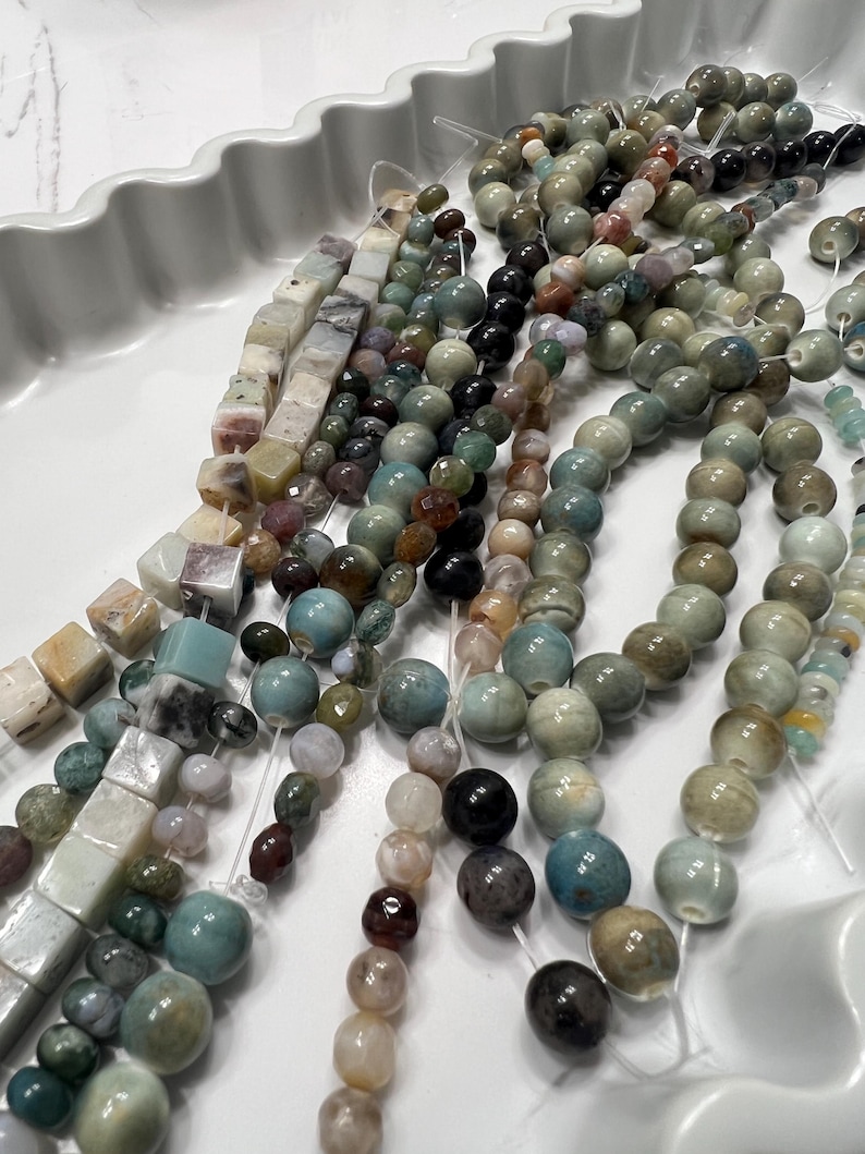 Puede incluir: Surtido de cuentas de piedras preciosas en varias formas y colores, incluyendo redondas, cuadradas e irregulares. Las cuentas est&aacute;n ensartadas, mostrando una paleta de verdes, azules, marrones y cremas. Las cuentas se exhiben en una bandeja blanca con bordes ondulados.