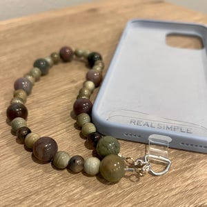 Puede incluir: Un colgante de tel&eacute;fono con cuentas en tonos tierra, incluyendo marrones, verdes y crema. El colgante est&aacute; unido a una funda de tel&eacute;fono azul claro con el texto "REAL SIMPLE".