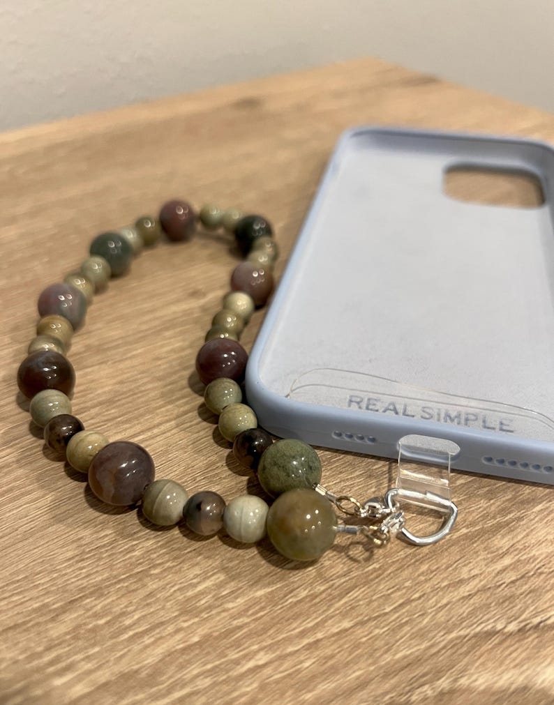 Puede incluir: Un colgante de tel&eacute;fono con cuentas en tonos tierra, incluyendo marrones, verdes y crema. El colgante est&aacute; unido a una funda de tel&eacute;fono azul claro con el texto "REAL SIMPLE".