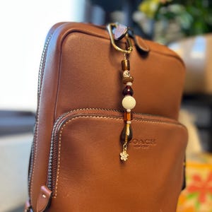 Puede incluir: Una mochila de cuero marr&oacute;n con un charm de cuentas. El charm presenta cuentas marrones, blancas y doradas, y un peque&ntilde;o charm de flor. La mochila tiene un bolsillo con cremallera y la marca "COACH NEW YORK" es visible.