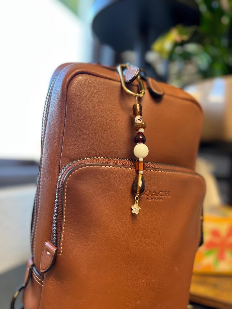 Puede incluir: Una mochila de cuero marr&oacute;n con un charm de cuentas. El charm presenta cuentas marrones, blancas y doradas, y un peque&ntilde;o charm de flor. La mochila tiene un bolsillo con cremallera y la marca "COACH NEW YORK" es visible.
