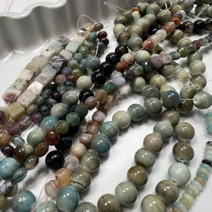 Puede incluir: Surtido de cuentas de piedras preciosas en varias formas y colores, incluyendo redondas, cuadradas y facetadas. Las cuentas son en tonos verdes, marrones, azules y negros, ensartadas en un hilo transparente. Las cuentas se muestran en una bandeja blanca con bordes.