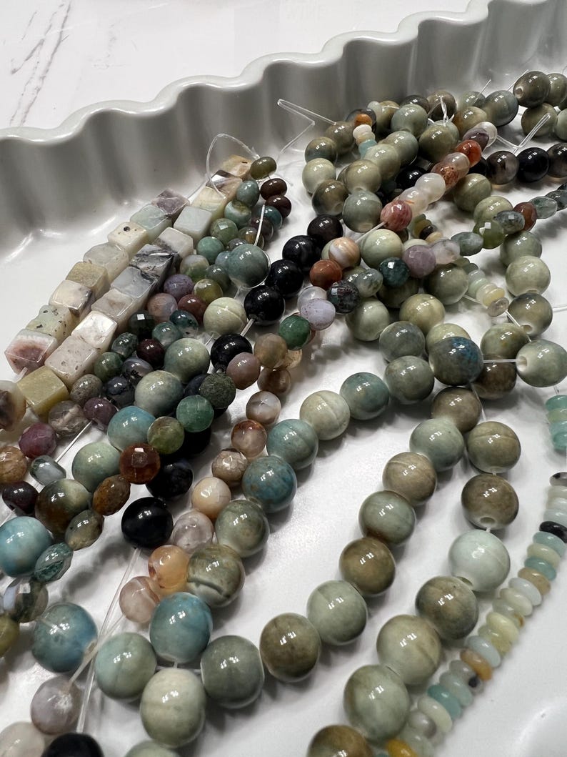 Puede incluir: Surtido de cuentas de piedras preciosas en varias formas y colores, incluyendo redondas, cuadradas y facetadas. Las cuentas son en tonos verdes, marrones, azules y negros, ensartadas en un hilo transparente. Las cuentas se muestran en una bandeja blanca con bordes.
