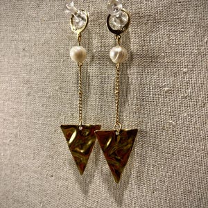 May include: A pair of gold-colored earrings with a pearl and a triangle pendant. The earrings have a gold chain and a clear, round stud. The triangle pendant has a textured surface.