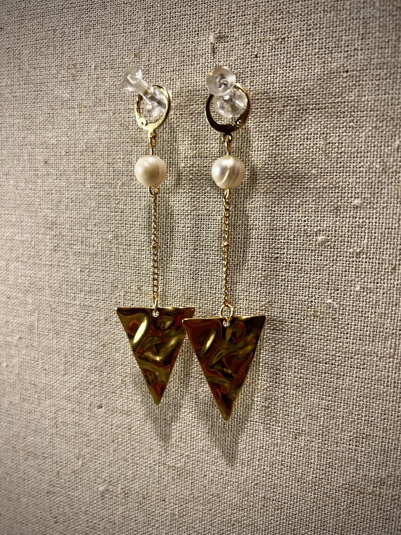 May include: A pair of gold-colored earrings with a pearl and a triangle pendant. The earrings have a gold chain and a clear, round stud. The triangle pendant has a textured surface.