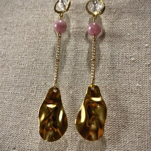 May include: A pair of gold-colored earrings with a unique design. Each earring features a clear stud, a gold hoop, a pink bead, a gold chain, and a textured, teardrop-shaped gold pendant. The earrings are displayed against a neutral-toned fabric background.