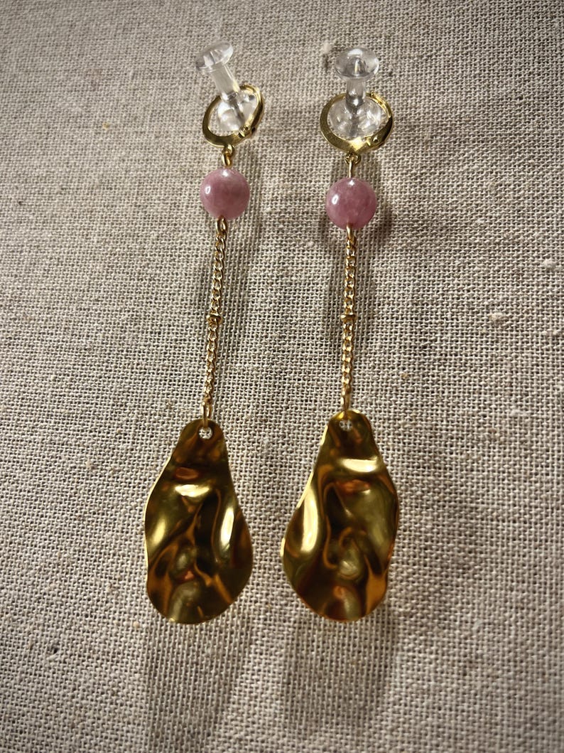 May include: A pair of gold-colored earrings with a unique design. Each earring features a clear stud, a gold hoop, a pink bead, a gold chain, and a textured, teardrop-shaped gold pendant. The earrings are displayed against a neutral-toned fabric background.