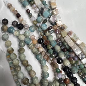 Puede incluir: Hebras de cuentas de piedras preciosas en varias formas y colores. Las cuentas incluyen estilos redondos, cuadrados y facetados en tonos de azul, verde, marr&oacute;n y crema. Las cuentas est&aacute;n ensartadas en una cuerda transparente.