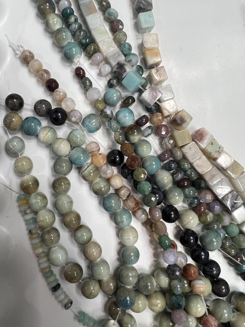 Puede incluir: Hebras de cuentas de piedras preciosas en varias formas y colores. Las cuentas incluyen estilos redondos, cuadrados y facetados en tonos de azul, verde, marr&oacute;n y crema. Las cuentas est&aacute;n ensartadas en una cuerda transparente.