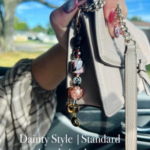 Puede incluir: Un llavero de cuentas con una variedad de cuentas coloridas, incluyendo negro, rosa y marr&oacute;n. El llavero est&aacute; unido a un bolso gris claro con una correa de cadena plateada. El texto "Dainty Style | Standard keychain clasp" es visible.