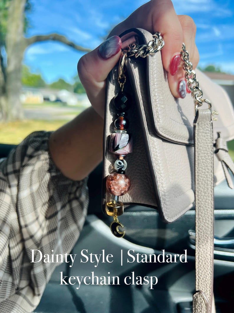 Puede incluir: Un llavero de cuentas con una variedad de cuentas coloridas, incluyendo negro, rosa y marr&oacute;n. El llavero est&aacute; unido a un bolso gris claro con una correa de cadena plateada. El texto "Dainty Style | Standard keychain clasp" es visible.