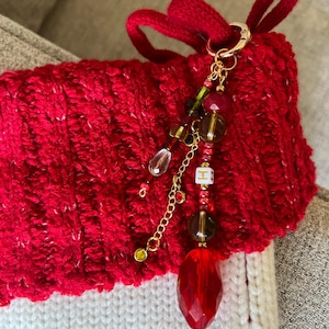 K&ouml;nnte beinhalten: Ein rot-wei&szlig; gestrickter Weihnachtsstrumpf mit einem goldfarbenen Anh&auml;nger. Der Anh&auml;nger hat eine Vielzahl von roten, gr&uuml;nen und klaren Perlen mit einem gro&szlig;en facettierten roten Kristall am unteren Ende. Ein kleiner wei&szlig;er W&uuml;rfel mit dem Buchstaben "H" ist ebenfalls enthalten.