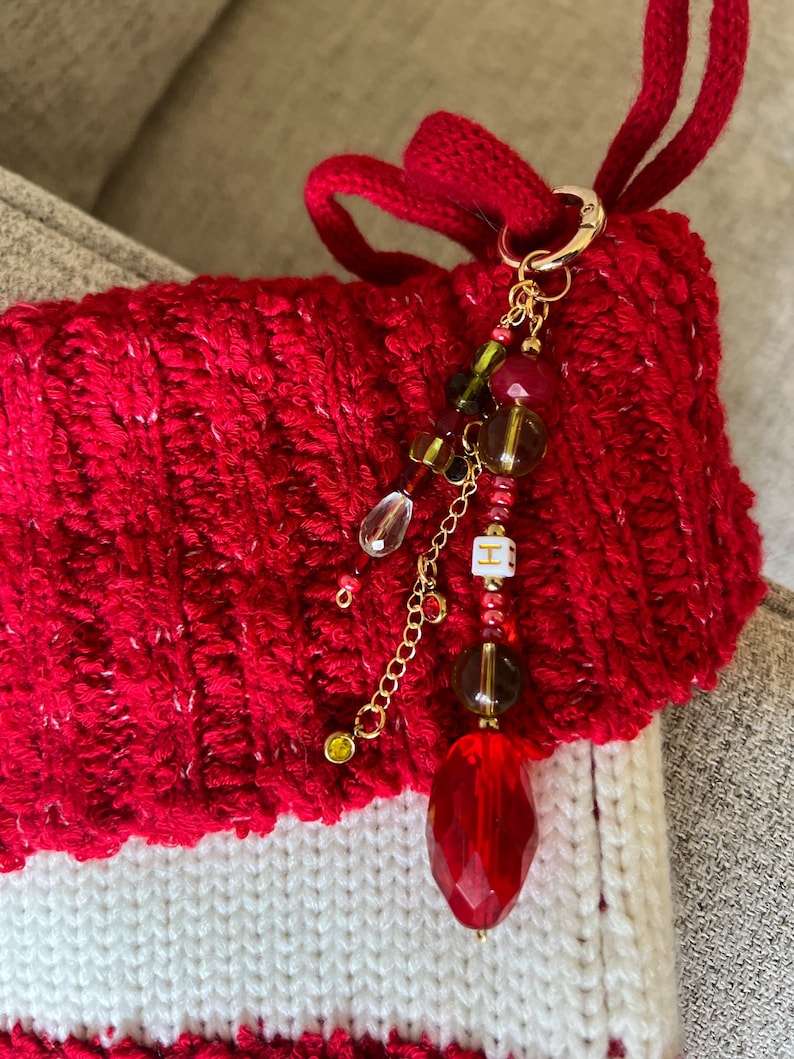 K&ouml;nnte beinhalten: Ein rot-wei&szlig; gestrickter Weihnachtsstrumpf mit einem goldfarbenen Anh&auml;nger. Der Anh&auml;nger hat eine Vielzahl von roten, gr&uuml;nen und klaren Perlen mit einem gro&szlig;en facettierten roten Kristall am unteren Ende. Ein kleiner wei&szlig;er W&uuml;rfel mit dem Buchstaben "H" ist ebenfalls enthalten.