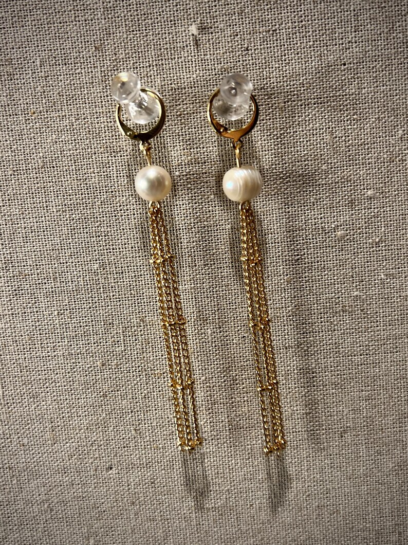 May include: A pair of gold-coloured drop earrings. Each earring has a clear stud, a gold hoop, a white pearl, and multiple strands of delicate gold chains. The earrings are set against a neutral, textured background.