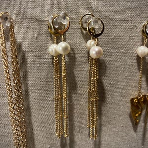 May include: Gold earrings featuring clear crystal accents, paired with dangling pearls and delicate chains. The earrings are presented against a neutral fabric backdrop, highlighting their elegant design. The jewelry is suitable for special occasions.