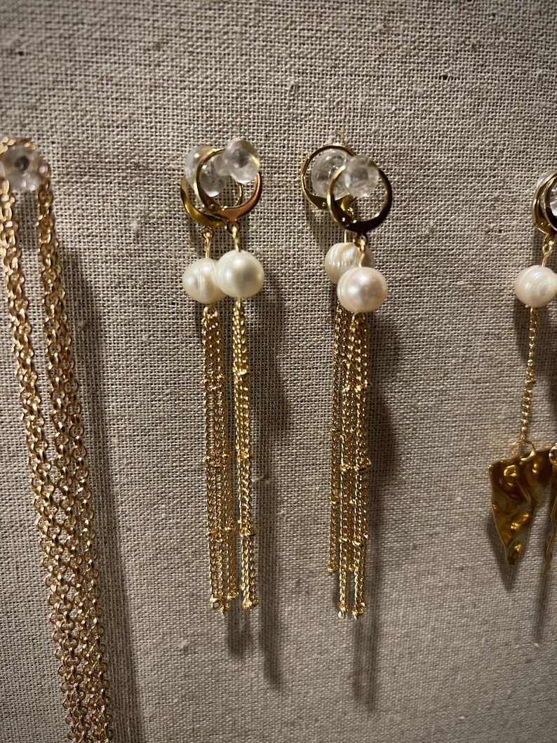 May include: Gold earrings featuring clear crystal accents, paired with dangling pearls and delicate chains. The earrings are presented against a neutral fabric backdrop, highlighting their elegant design. The jewelry is suitable for special occasions.