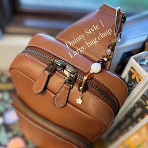Puede incluir: Un bolso de cuero marr&oacute;n con cierre de cremallera y un cierre de cuentas decorativo. El cierre presenta un peque&ntilde;o dije de flor y varias cuentas en tonos marr&oacute;n, blanco y dorado. El texto "Dainty Style | Large bag clasp" es visible.