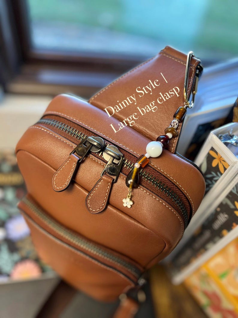 Puede incluir: Un bolso de cuero marr&oacute;n con cierre de cremallera y un cierre de cuentas decorativo. El cierre presenta un peque&ntilde;o dije de flor y varias cuentas en tonos marr&oacute;n, blanco y dorado. El texto "Dainty Style | Large bag clasp" es visible.