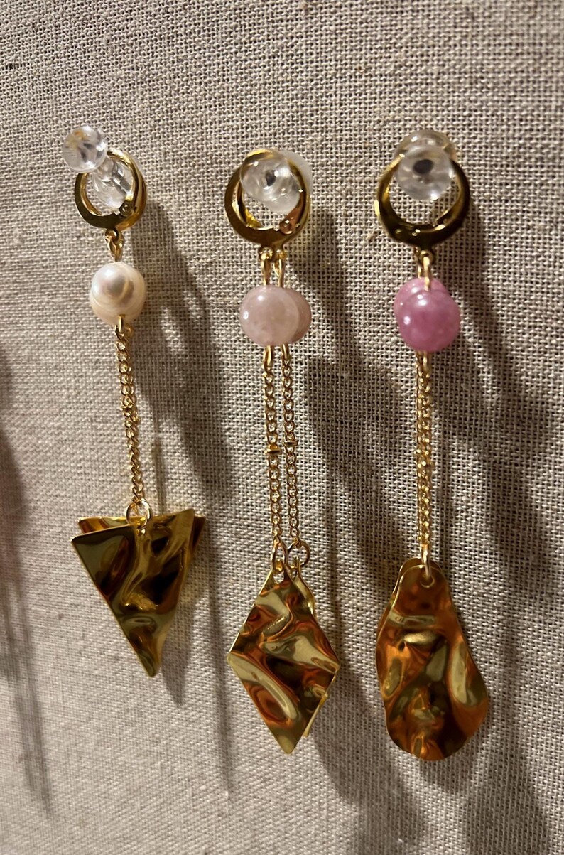 May include: Three pairs of gold-toned earrings with different designs. Each earring features a clear, round stud, a chain, and a unique charm: a pearl and triangle, a pink stone and diamond, and a pink stone and teardrop shape. The earrings hang against a neutral fabric.