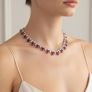 May include: A close-up of a necklace featuring alternating oval-cut red gemstones and round, clear gemstones. The necklace is worn on a person wearing a white spaghetti strap dress.