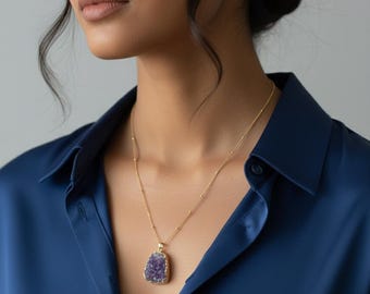 May include: A gold necklace with a pendant featuring a large, irregular-shaped, purple amethyst stone. The necklace is worn over a dark blue collared shirt.