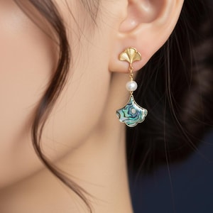 May include: Gold-toned earrings featuring a ginkgo leaf design. Each earring has a small pearl and a fan-shaped pendant with iridescent abalone shell inlay. The earrings are dangling and worn on an ear.