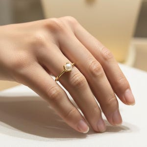 May include: A gold ring featuring a small, round pearl. The ring is set on a delicate gold band, showcasing the pearl as the focal point. The ring is on a person's finger.