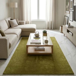 May include: A modern living room with a beige sectional sofa, a wooden coffee table with a glass top, and a long, olive green rug. The room has a large window with sheer curtains and a marble floor.